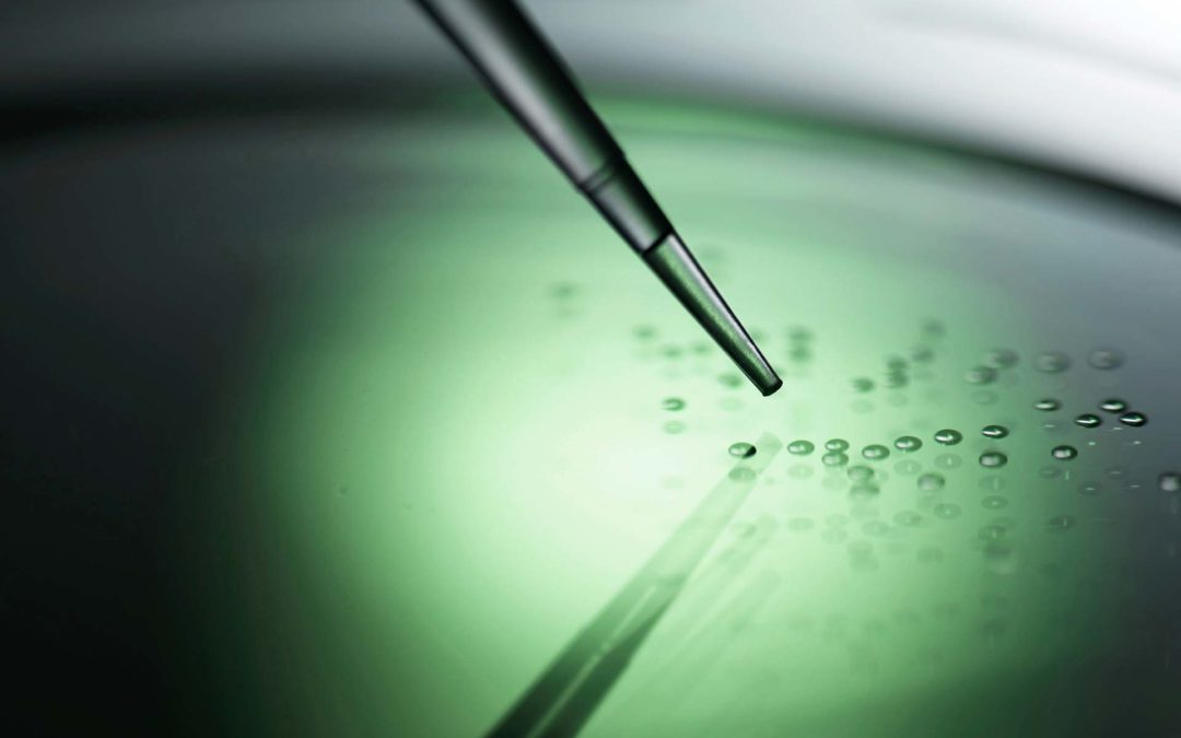 The tip of a pipette pointing towards droplets suspended in a green solution.